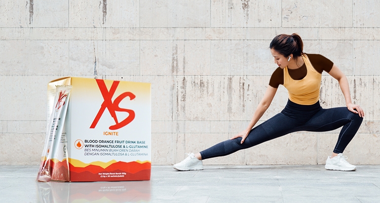 Image of XS IGNITE Blood Orange Fruit Drink Base With Isomaltulose & L-Glutamine next to a healthy woman in sportswear doing stretches to warm up before exercise 